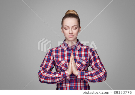 Portrait of calm beautiful blonde girl in red, pink checkered shirt, collected bun hairstyle, makeup standing, closed eyes and yoga meditation hands. indoor studio shot. isolated on gray background Portrait of calm beautiful blonde girl in red, pink checkered shirt, collected bun hairstyle, makeup standing, closed eyes and yoga meditation hands. indoor studio shot. isolated on gray background 89353776