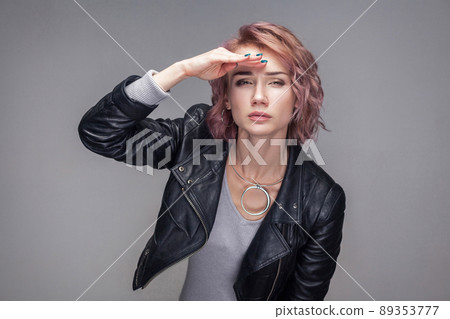 Portrait of attentive beautiful girl with short hairstyle, makeup in casual style black leather jacket standing with hand on forehead and looking far. indoor studio shot, isolated on grey background. 89353777
