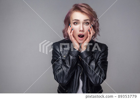Portrait of shocked girl with short hairstyle, makeup in black leather jacket standing touching face and looking at camera with big eyes and open mouth. indoor studio shot, isolated on grey background Portrait of shocked girl with short hairstyle, makeup in black leather jacket standing touching face and looking at camera with big eyes and open mouth. indoor studio shot, isolated on grey background 89353779