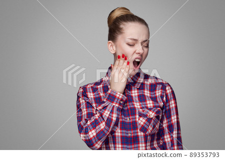 Tooth pain. unhappy blonde girl in red, pink checkered shirt, collected bun hairstyle, makeup standing touching her painful tooth with open mouth. indoor studio shot. isolated on gray background Tooth pain. unhappy blonde girl in red, pink checkered shirt, collected bun hairstyle, makeup standing touching her painful tooth with open mouth. indoor studio shot. isolated on gray background 89353793