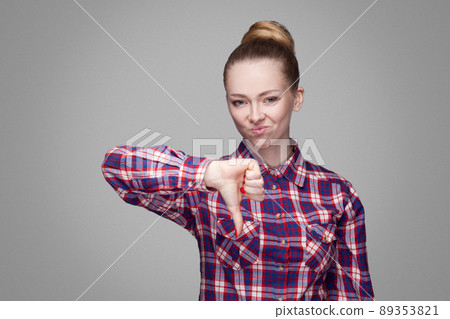 Unhappy sad girl with pink checkered shirt, collected bun hairstyle and makeup standing and looking at camera with unsatisfied face and thumbs down. indoor studio shot, isolated on gray background. Unhappy sad girl with pink checkered shirt, collected bun hairstyle and makeup standing and looking at camera with unsatisfied face and thumbs down. indoor studio shot, isolated on gray background. 89353821