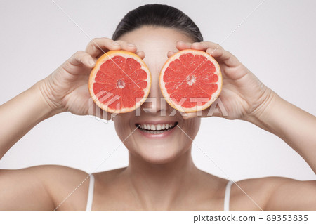 Closeup portrait of happy woman with perfect skin smiling after cream, balm, mask, lotion, holding half of grapefruit and covering her eye, isolated on grey background. Indoor, studio shot,copy space 89353835
