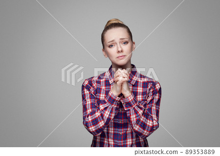 Please help me. beautiful blonde girl in red, pink checkered shirt, collected bun hairstyle standing and looking at camera with palm hands and begging. indoor studio shot. isolated on gray background 89353889