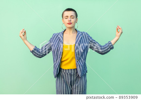 Portrait of calm handsome beautiful short hair young stylish woman in casual striped suit standing in yoga pose, closed eyes and relaxing. indoor studio shot isolated on light green background. Portrait of calm handsome beautiful short hair young stylish woman in casual striped suit standing in yoga pose, closed eyes and relaxing. indoor studio shot isolated on light green background. 89353909