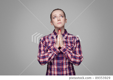 Prayer beautiful blonde girl in red, pink checkered shirt, collected bun hairstyle, makeup standing and looking to up and pleading or begging. indoor studio shot. isolated on gray background 89353929
