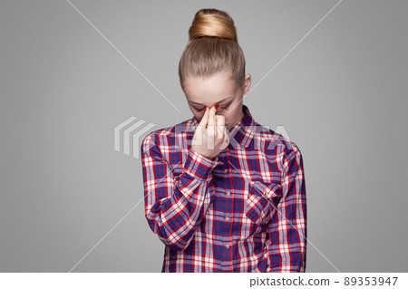 Sad, tired or depressed beautiful blonde girl in red, pink checkered shirt, collected bun hairstyle, makeup standing and holding head down and crying. indoor studio shot. isolated on gray background 89353947