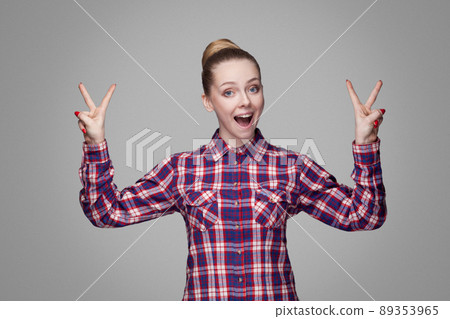 Beautiful girl with pink checkered shirt, collected bun hairstyle and makeup standing and showing victory or peace sign, looking at camera with toothy smile. indoor shot isolated on gray background 89353965