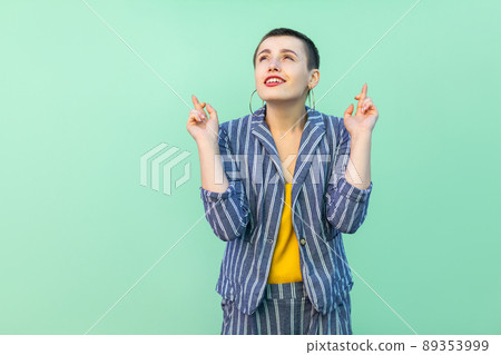 Portrait of hopeful handsome beautiful short hair young stylish woman in casual striped suit standing, crossed fingers and wishful to win . indoor studio shot isolated on light green background. Portrait of hopeful handsome beautiful short hair young stylish woman in casual striped suit standing, crossed fingers and wishful to win . indoor studio shot isolated on light green background. 89353999