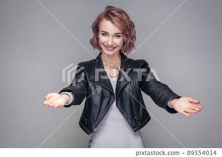 Portrait of happy girl with short hair, makeup in casual style black leather jacket standing with raised arms, toothy smile, hug, generosity or welcome. indoor studio shot, isolated on grey background 89354014
