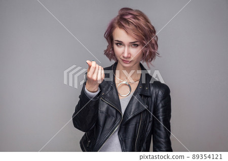 Portrait of funny beautiful girl with short hairstyle and makeup in casual style black leather jacket standing with money gesture and looking at camera. indoor studio shot, isolated on grey background Portrait of funny beautiful girl with short hairstyle and makeup in casual style black leather jacket standing with money gesture and looking at camera. indoor studio shot, isolated on grey background 89354121
