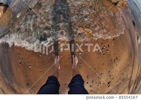 First Person Fisheye Perspective of Man's Feet in Shallow Water with Waves Rolling into Sandy Beach 89354167