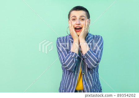 Portrait of surprised beautiful short hair young stylish woman in casual striped suit standing touching her face and looking at camera with shocked face. studio shot isolated on light green background 89354202