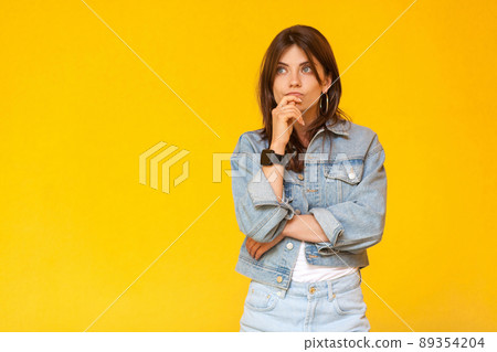 Portrait of thoughtful beautiful brunette young woman with makeup in denim casual style standing, touching her lips, looking away and thinking what to do. studio shot, isolated on yellow background. 89354204