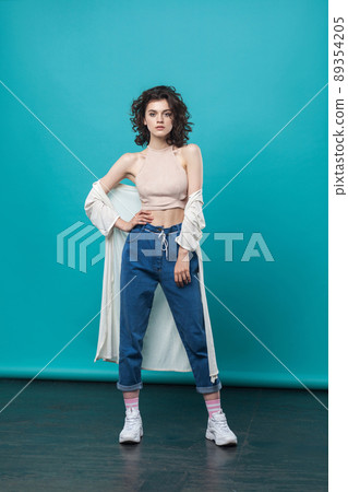 full length of beautiful young brunette woman in casual style standing and posing against blue wall. looking at camera with serious face. indoor studio shot. 89354205