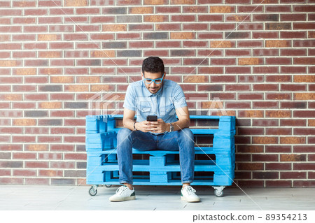 Full length portrait of successful handsome young bearded man in casual style, eyeglasses sitting on blue wooden pallet, holding, looking at mobile smartphone. indoor studio shot on brown brick wall. Full length portrait of successful handsome young bearded man in casual style, eyeglasses sitting on blue wooden pallet, holding, looking at mobile smartphone. indoor studio shot on brown brick wall. 89354213