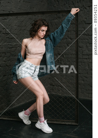 Full length picture of beautiful young woman with makeup and in casual style near fance standing and posing on dark brick wall background and looking away. indoor studio shot. 89354251