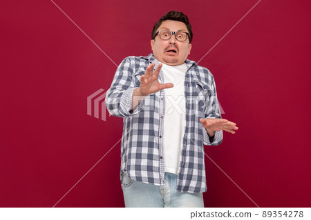 Portrait of shocked handsome middle aged business man in casual checkered shirt and eyeglasses standing and looking at camera with scared eyes. indoor studio shot, isolated on dark red background. 89354278