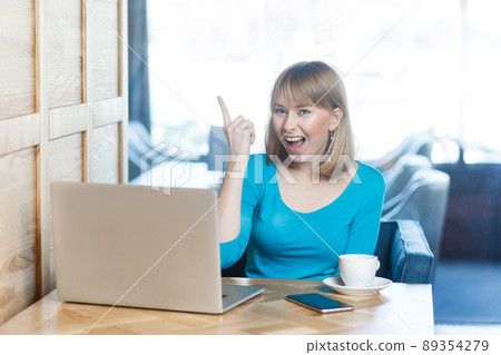 Have idea! Portrait of creative positive attractive young girl freelancer in blue t-shirt are sitting in cafe and working on laptop with toothy smile and showing finger up, looking at camera. indoor Have idea! Portrait of creative positive attractive young girl freelancer in blue t-shirt are sitting in cafe and working on laptop with toothy smile and showing finger up, looking at camera. indoor 89354279