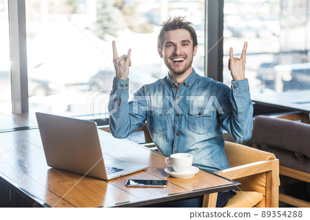 Portrait of handsome successfull positive bearded young freelancer in blue jeans shirt are sitting in cafe and working on laptop with toothy smile and showing rock sign, looking at camera. indoor Portrait of handsome successfull positive bearded young freelancer in blue jeans shirt are sitting in cafe and working on laptop with toothy smile and showing rock sign, looking at camera. indoor 89354288