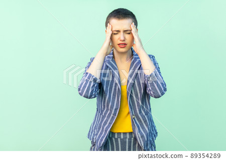 Headache or confusion. Portrait of handsome beautiful short hair young woman in casual striped suit standing holding her painful head or thinking. studio shot isolated on light green background. Headache or confusion. Portrait of handsome beautiful short hair young woman in casual striped suit standing holding her painful head or thinking. studio shot isolated on light green background. 89354289
