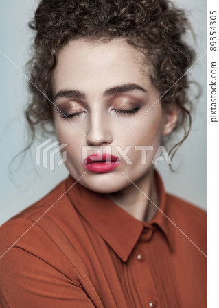 Beauty portrait of young beautiful fashion calm relaxed model with collected dark curly hair, nude makeup, red lips, and brown shirt and closed eyes. indoor studio shot. isolated on gray background. Beauty portrait of young beautiful fashion calm relaxed model with collected dark curly hair, nude makeup, red lips, and brown shirt and closed eyes. indoor studio shot. isolated on gray background. 89354305