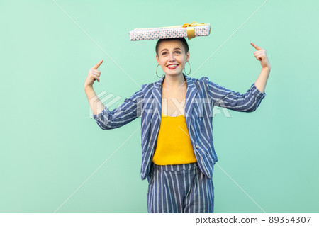 Portrait of playful beautiful with short hair young woman in striped suit standing, holding present box on head and pointing fingers to it, toothy smile. Indoor, isolated, studio shot,green background Portrait of playful beautiful with short hair young woman in striped suit standing, holding present box on head and pointing fingers to it, toothy smile. Indoor, isolated, studio shot,green background 89354307