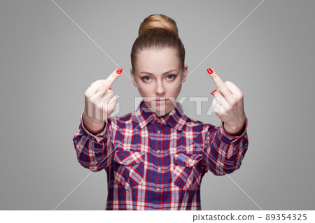 angry beautiful blonde girl in pink checkered shirt and collected bun hairstyle standing, looking at camera and showing middle finger with poker face. indoor studio shot. isolated on gray background 89354325