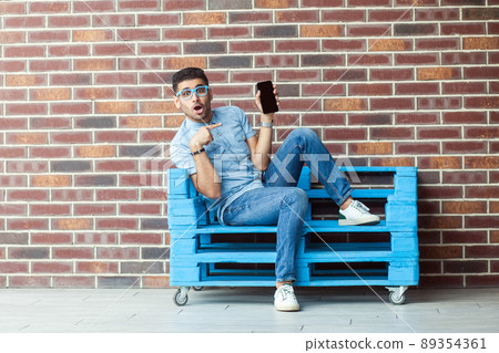 Full length portrait of shocked young bearded man in casual style and eyeglasses sitting on blue wooden pallet and pointing at mobile screen, looking at camera. indoor studio shot on brown brick wall. Full length portrait of shocked young bearded man in casual style and eyeglasses sitting on blue wooden pallet and pointing at mobile screen, looking at camera. indoor studio shot on brown brick wall. 89354361