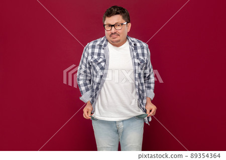 I don't have money more. Portrait of poor sad middle aged man in casual checkered shirt and eyeglasses standing looking and showing his empty pocket. indoor studio shot isolated on dark red background 89354364