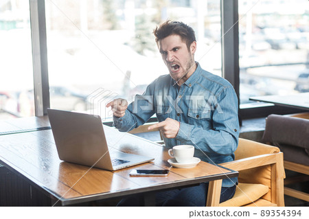 Yes, it`s you! Side view portrait of angry upset bearded young big boss in blue jeans shirt are sitting in cafe, working and making video call on laptop, pointing finger and blame the companion. Yes, it`s you! Side view portrait of angry upset bearded young big boss in blue jeans shirt are sitting in cafe, working and making video call on laptop, pointing finger and blame the companion. 89354394