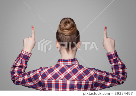 back view of beautiful blonde girl in red, pink checkered shirt, collected bun hairstyle standing and showing up with fingers. indoor studio shot. isolated on gray background back view of beautiful blonde girl in red, pink checkered shirt, collected bun hairstyle standing and showing up with fingers. indoor studio shot. isolated on gray background 89354497