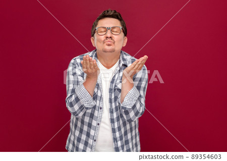 Portrait of happy in love handsome middle aged business man in casual checkered shirt, eyeglasses standing, closed eyes and sending kiss at camera. indoor studio shot, isolated on dark red background. Portrait of happy in love handsome middle aged business man in casual checkered shirt, eyeglasses standing, closed eyes and sending kiss at camera. indoor studio shot, isolated on dark red background. 89354603