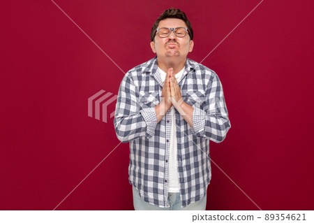 Please help me. Portrait of worried middle aged man in checkered shirt, eyeglasses standing with palm hands pleading, hopeful for something or forgiveness. studio shot, isolated on dark red background Please help me. Portrait of worried middle aged man in checkered shirt, eyeglasses standing with palm hands pleading, hopeful for something or forgiveness. studio shot, isolated on dark red background 89354621