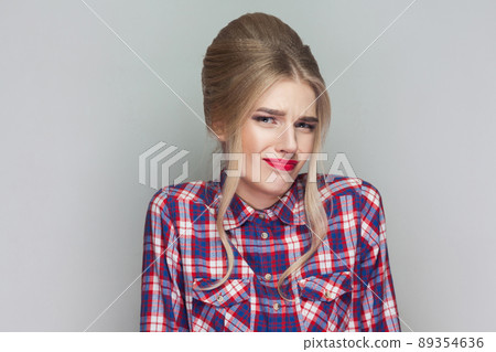I dont know. confused beautiful girl with pink checkered shirt, collected updo hairstyle and makeup standing and looking away with doubtful face. indoor studio shot, isolated on gray background. 89354636