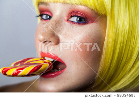 Beauty portrait of cute young female model with freckles, red makeup and yellow wig, holding and bitting her colorful candy stick and looking at camera. indoor studio shot, isolated on gray background Beauty portrait of cute young female model with freckles, red makeup and yellow wig, holding and bitting her colorful candy stick and looking at camera. indoor studio shot, isolated on gray background 89354695
