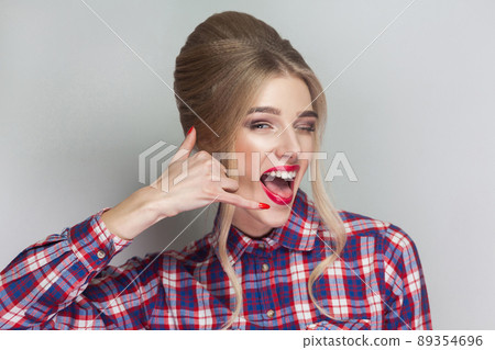 Call me! funny beautiful blonde girl in pink checkered shirt, collected updo hairstyle, red lips and makeup standing and looking at camera with calling gesture. indoor, isolated on gray background Call me! funny beautiful blonde girl in pink checkered shirt, collected updo hairstyle, red lips and makeup standing and looking at camera with calling gesture. indoor, isolated on gray background 89354696