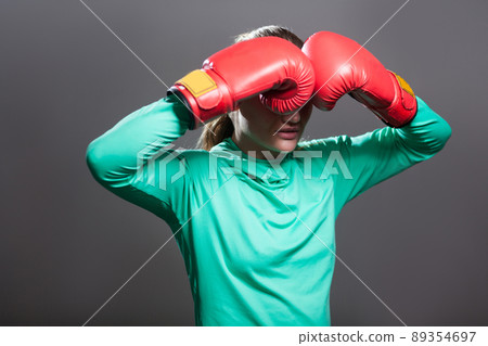 Sad young athlete woman with collected hair standing in position, holding hands in boxing red gloves near eyes and hiding tears after knockout. Indoor studio shot, isolated on dark grey background. Sad young athlete woman with collected hair standing in position, holding hands in boxing red gloves near eyes and hiding tears after knockout. Indoor studio shot, isolated on dark grey background. 89354697