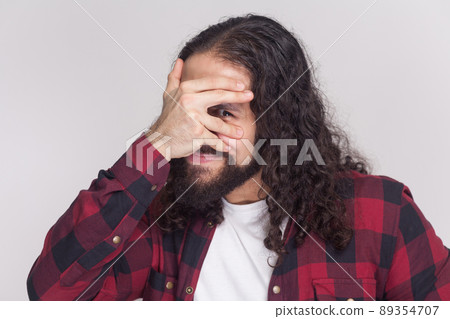 Portrait of handsome funny man with beard and black long curly hair in casual style, checkered red shirt standing hiding face and looking peek. indoor studio shot, isolated on grey background. 89354707