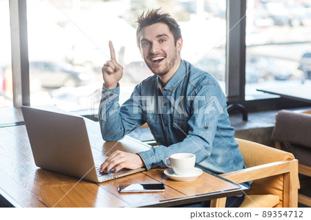 Have idea! Portrait of creative positive bearded young freelancer in blue jeans shirt are sitting in cafe and working on laptop with toothy smile and showing finger up, looking at camera. indoor Have idea! Portrait of creative positive bearded young freelancer in blue jeans shirt are sitting in cafe and working on laptop with toothy smile and showing finger up, looking at camera. indoor 89354712