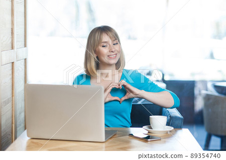 Love you! Portrait of romantic happy young girl with blonde hair in blue blouse are sitting in cafe and looking at display with a heart shape with fingers through a webcam online video call. Indoor 89354740