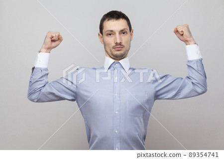 I am strong. Portrait of serious proud handsome bristle businessman in classic blue shirt standing and looking at camera with raised arms. indoor studio shot, isolated on grey background copyspace. I am strong. Portrait of serious proud handsome bristle businessman in classic blue shirt standing and looking at camera with raised arms. indoor studio shot, isolated on grey background copyspace. 89354741
