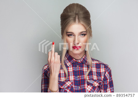angry beautiful blonde girl in pink checkered shirt and collected updo hairstyle standing, looking at camera and showing middle finger with poker face. indoor studio shot. isolated on gray background 89354757