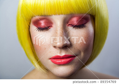 Beauty portrait of calm cute young female model with freckles, red makeup and yellow wig, closed eyes with relaxed serious face. indoor studio shot, isolated on gray background. Beauty portrait of calm cute young female model with freckles, red makeup and yellow wig, closed eyes with relaxed serious face. indoor studio shot, isolated on gray background. 89354769