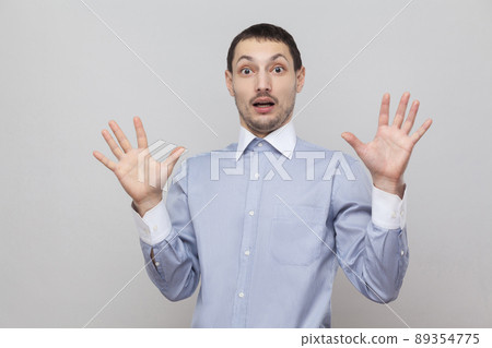 Portrait of shocked handsome bristle businessman in classic blue shirt standing with raised arms and looking at camera with scared face . indoor studio shot, isolated on grey background copyspace. Portrait of shocked handsome bristle businessman in classic blue shirt standing with raised arms and looking at camera with scared face . indoor studio shot, isolated on grey background copyspace. 89354775