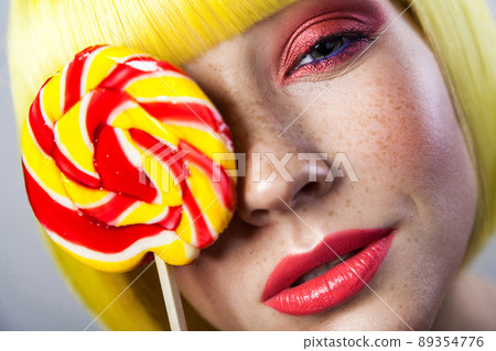 Beauty portrait of cute young girl with freckles, red makeup and yellow wig, holding colorful candy stick on eye and looking at camera with passion. indoor studio shot, isolated on gray background. Beauty portrait of cute young girl with freckles, red makeup and yellow wig, holding colorful candy stick on eye and looking at camera with passion. indoor studio shot, isolated on gray background. 89354776