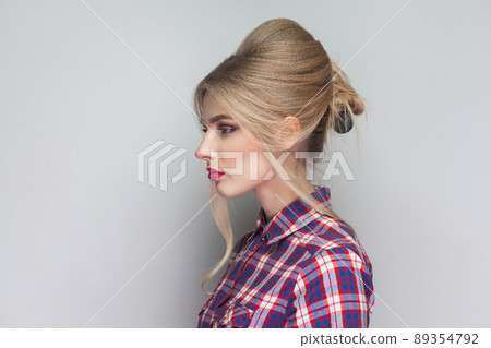 Profile side view of beautiful girl with pink checkered shirt, collected updo hairstyle, red lips and makeup standing with seious face and looking away. indoor studio shot, isolated on gray background Profile side view of beautiful girl with pink checkered shirt, collected updo hairstyle, red lips and makeup standing with seious face and looking away. indoor studio shot, isolated on gray background 89354792
