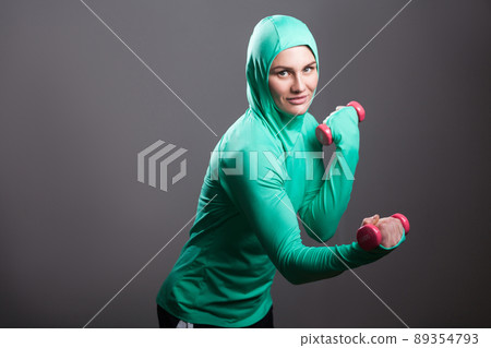 Beautiful muslim woman in green hijab or islamic sportswear standing, holding red dumbbells training biceps, looking at camera with toothy smile. indoor studio shot, isolated on dark grey background 89354793