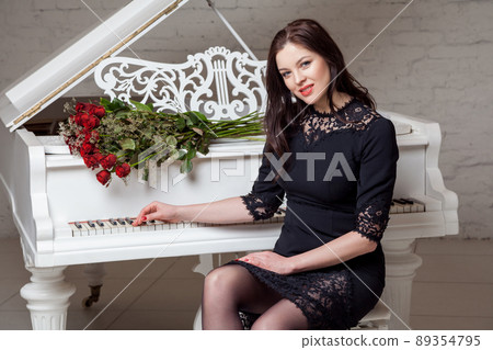 Beautiful happy brunette woman in black classic dress with bouquet of red roses sitting at piano and looking at camera with toothy smile. indoor studio shot on white brick wall. Beautiful happy brunette woman in black classic dress with bouquet of red roses sitting at piano and looking at camera with toothy smile. indoor studio shot on white brick wall. 89354795