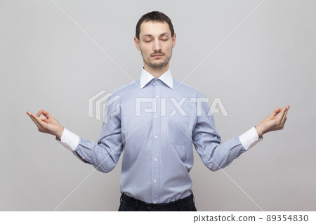 Portrait of calm handsome bristle businessman in classic light blue shirt standing in yoga pose with closed eyes and meditating. indoor studio shot, isolated on grey background copyspace. 89354830