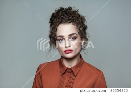 Beauty portrait of young beautiful fashion model with collected dark curly hair, nude makeup, red lips, and brown shirt looking at camera serious. indoor studio shot. isolated on gray background. 89354833